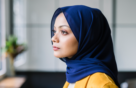 Woman wearing blue hijab standing by a window looking outside.