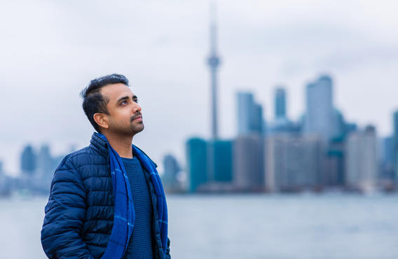 A man standing by a harbor in Toronto staring off into the distance with his hands in his pockets. Behind him is the landscape of downtown Toronto with the CN tower visibile.